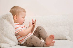 Adorable toddler boy sitting on the sofa in the living room and playing with smartphone. Child learning how to use smartphone. Boy texting on the phone. - technology and lifestyle concept.