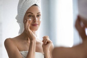 Attractive young adult woman applying facial cream looking in mirror