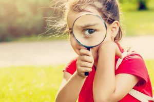 Positive cheerful little girl looking through a magnifying glass outdoors