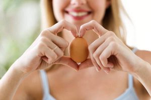 Beautiful young woman showing egg with hands in a heart shape at home.