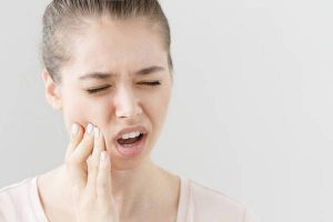 Close headshot of young European female isolated on grey background suffering from severe toothache, feeling pain so strong that she is pressing fingers to cheek to calm it down, looking desperate.