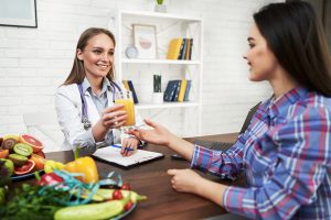 A smiling nutritionist advises a young patient woman on proper nutrition and dieting. The doctor shows a scheme of weight loss without a bad effect on health. Diet based on vegetables and fruits juice