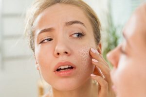 Young beautiful woman touching skin in bathroom. Unhappy girl standing in towel, looking in the mirror, checking dry irritated skin. Morning skincare routine.