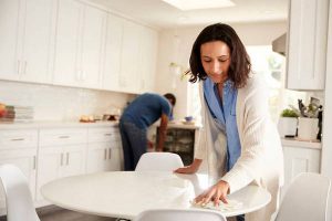 Millennial woman cleaning kitchen table while her partner also does chores in the background, focus on foreground