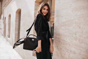 Joyful young brunette with fluffy hair, red lips, trendy dress and black jacket, belt on waist stan
