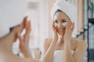 Attractive latina woman with under eye patches looking at mirror during daily skincare treatment